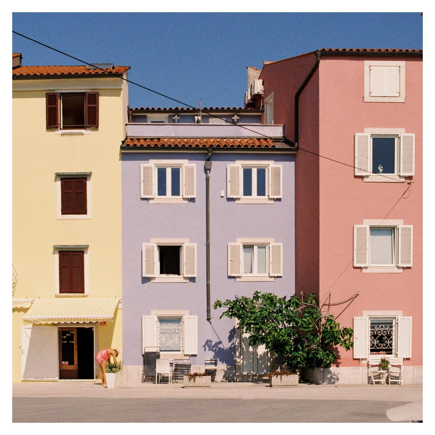 A woman tends to a plant in front of colorful buildings with white shutters under a clear blue sky.