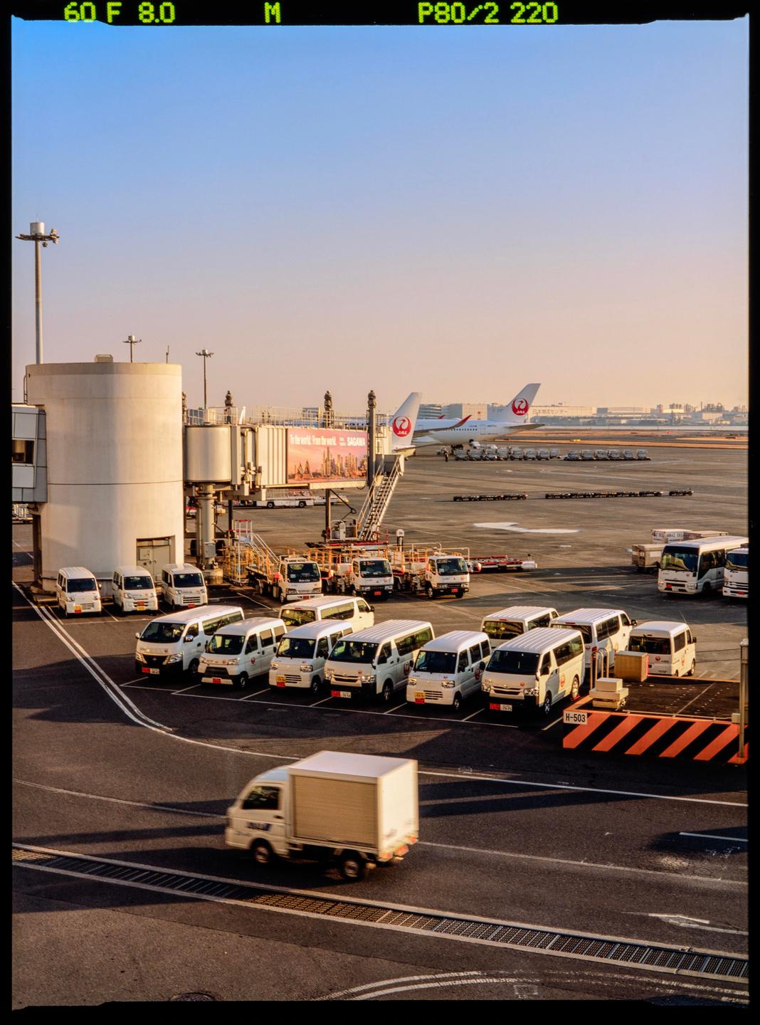 An airplane is parked at an airport gate with several vans and trucks on the tarmac during sunset.