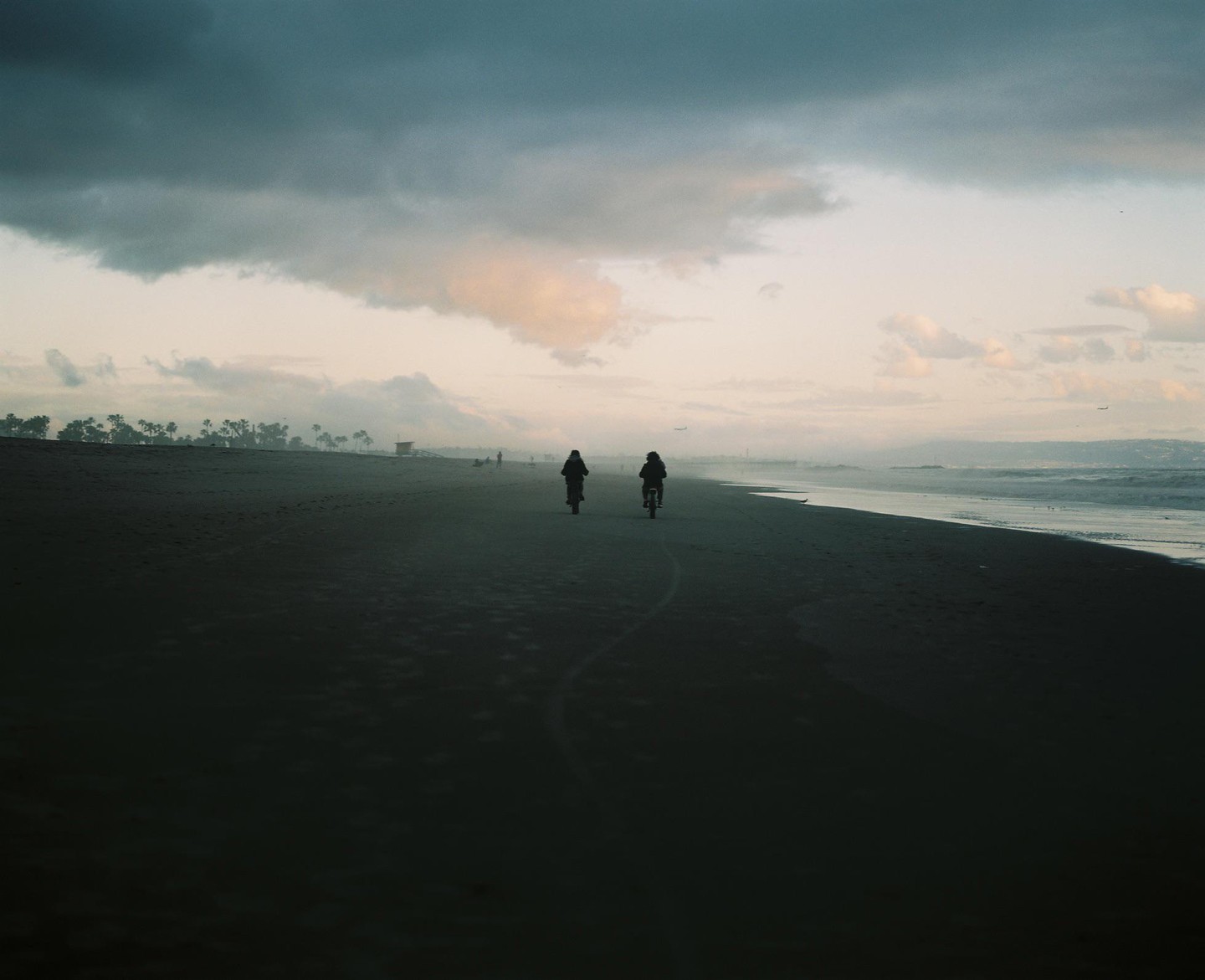Two people ride bicycles along a sandy beach next to the ocean under a cloudy sky.