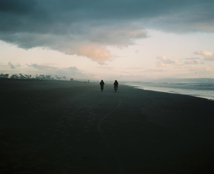 Two people ride bicycles along a sandy beach next to the ocean under a cloudy sky.