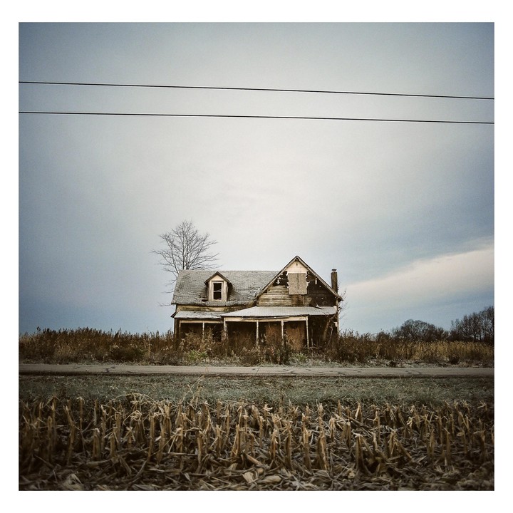 An abandoned house stands in a field under a cloudy sky.