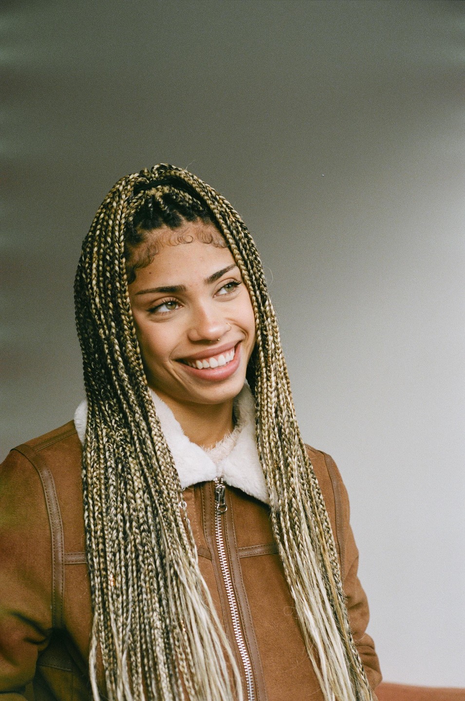 A woman with long braids and a brown jacket smiles while looking to the side.