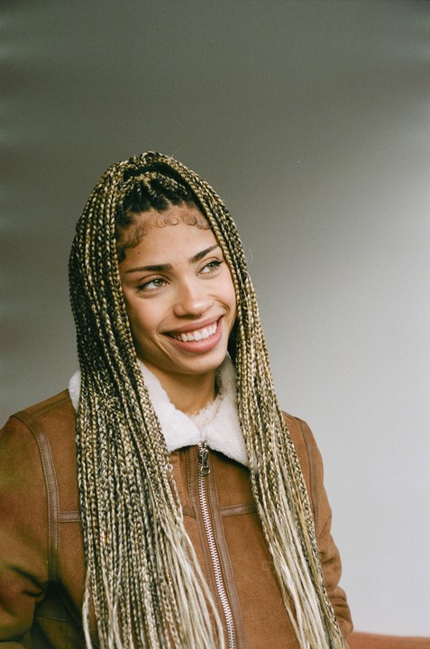 A woman with long braids and a brown jacket smiles while looking to the side.