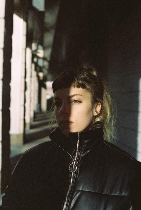 A woman with bangs and a ponytail wears a black puffer jacket, with half her face in shadow and half in sunlight.
