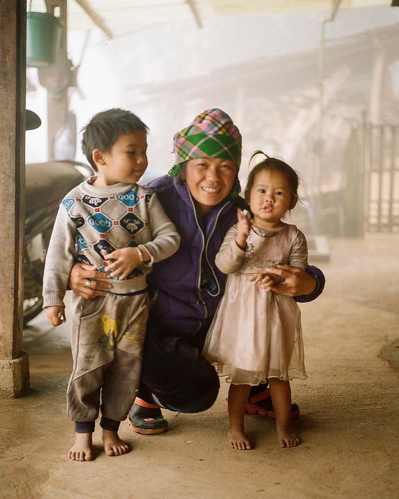 A woman wearing a plaid hat crouches between a young boy and a young girl, all three smiling at the camera.