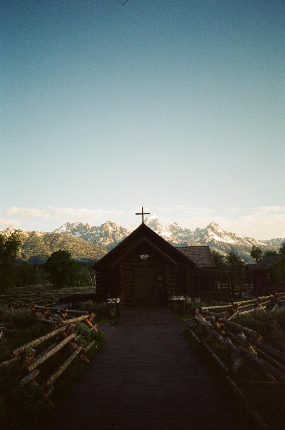 A wooden chapel with a cross on its roof stands before a mountain range with snow-capped peaks.