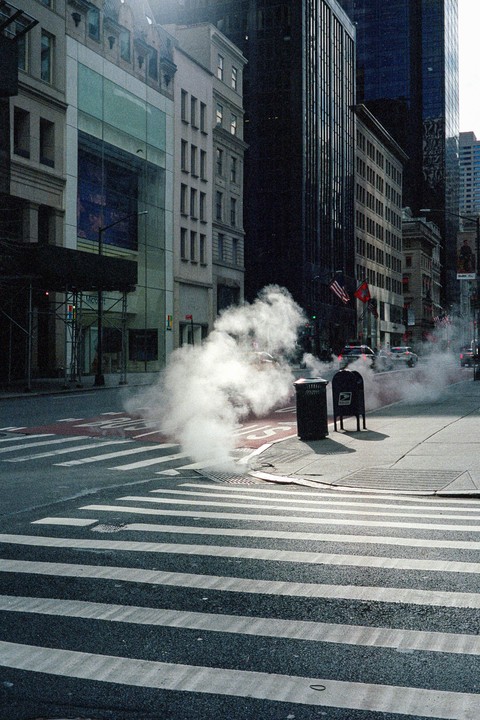 Steam rises from a manhole cover on a city street with tall buildings and a crosswalk.