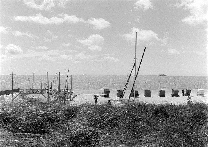 A beach with tall grass in the foreground, several beach chairs, a boat, and a person walking towards the water.