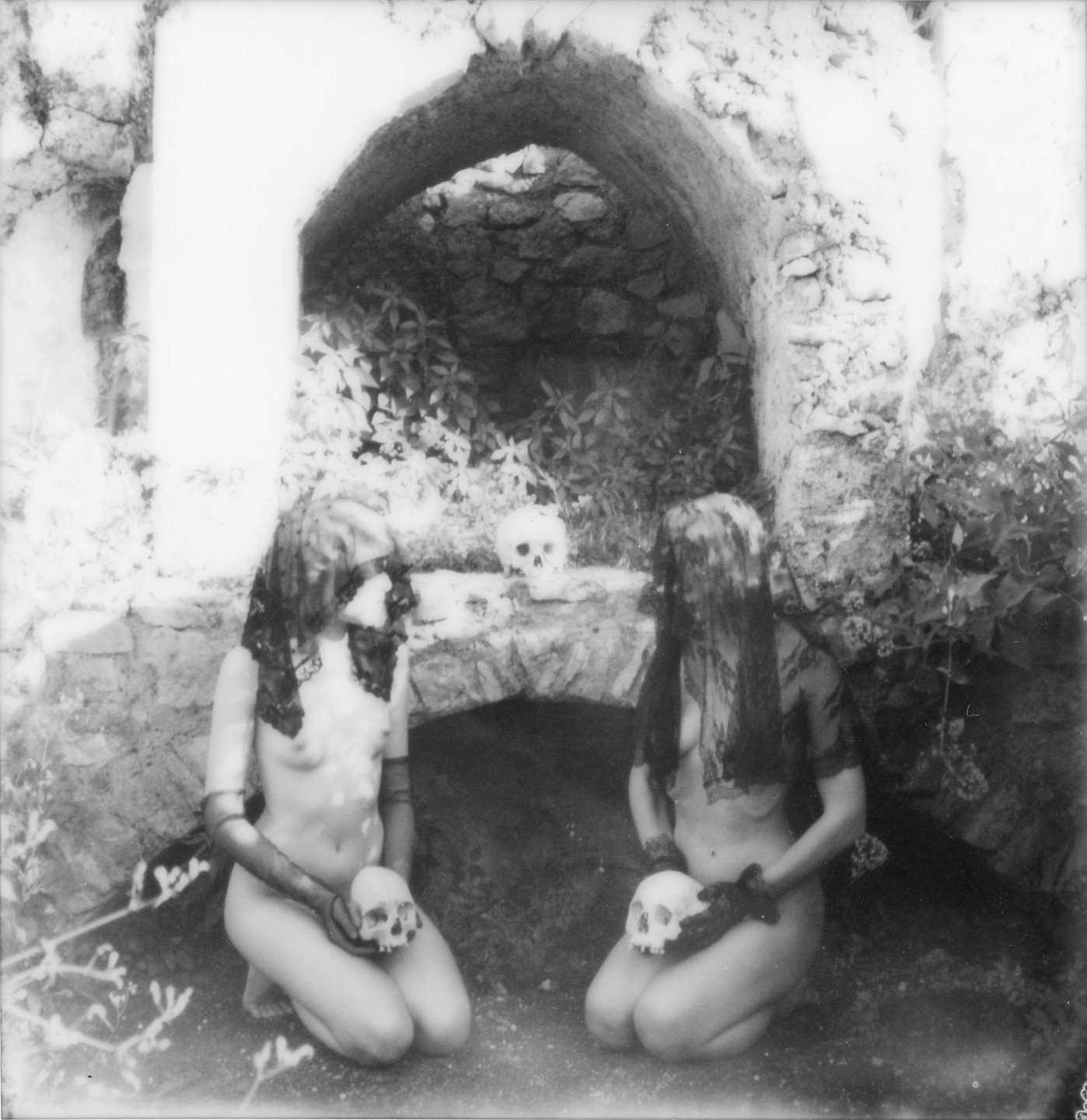 Two women kneel in front of a stone archway, each holding a skull, with another skull resting on the archway.