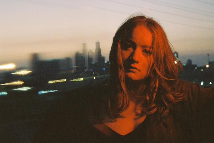 A woman with long hair is illuminated by orange light with a blurry city skyline in the background.