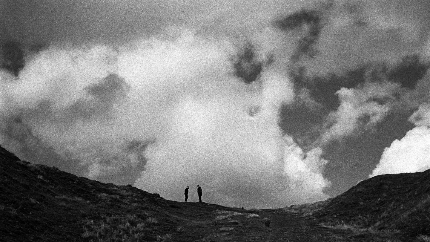 Two people stand on a grassy hill under a dramatic cloudy sky.