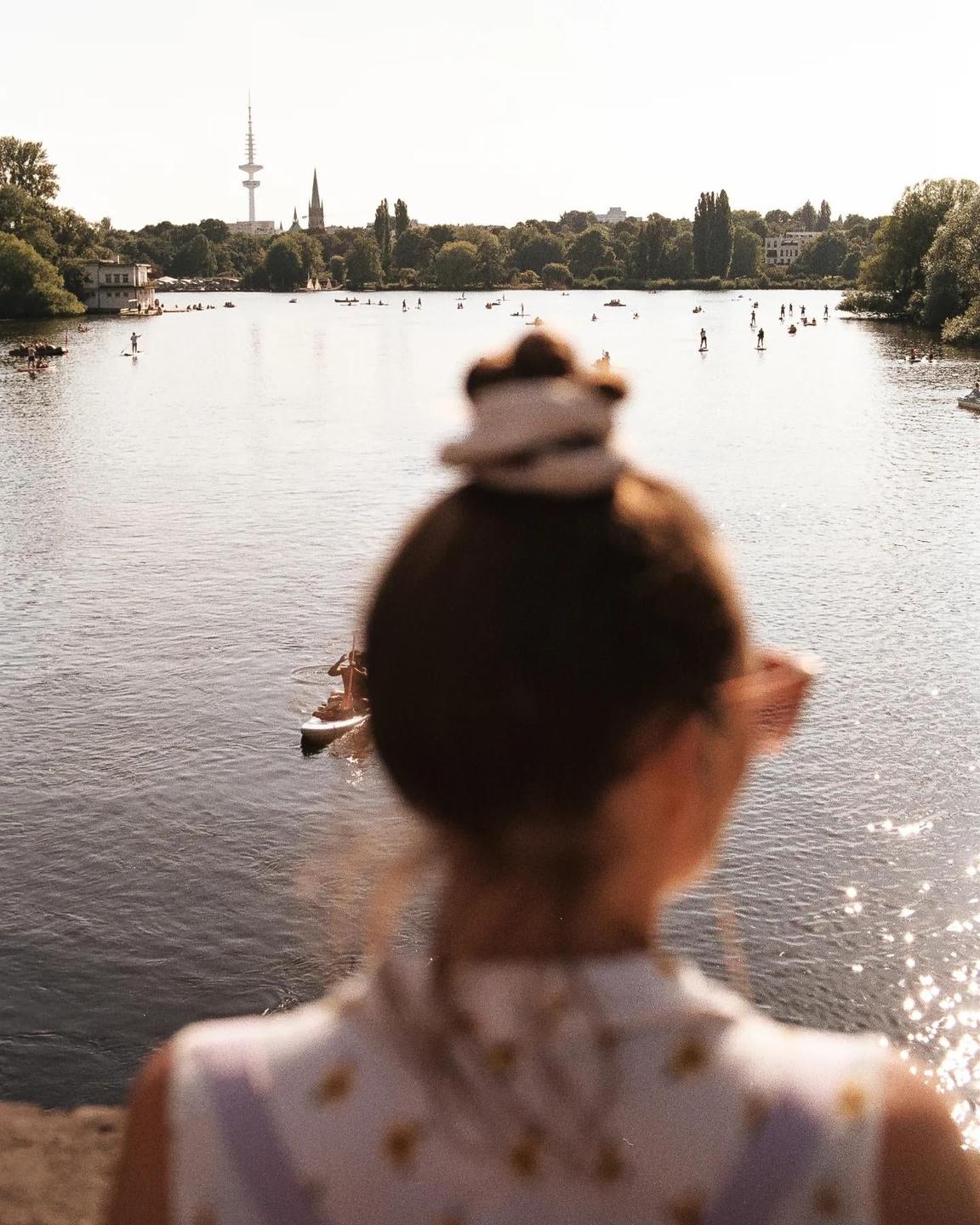 A woman with her back to the viewer looks out over a lake with many paddleboarders and a distant city skyline.