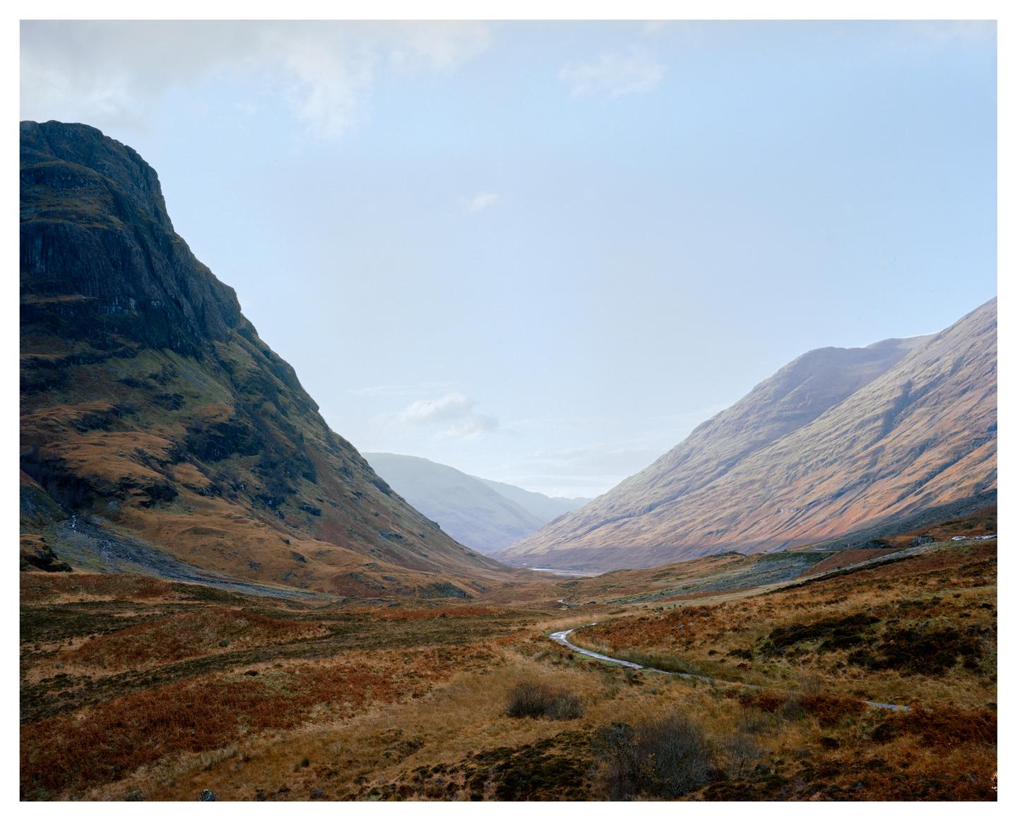 A winding road leads through a valley between two large mountains under a bright sky.