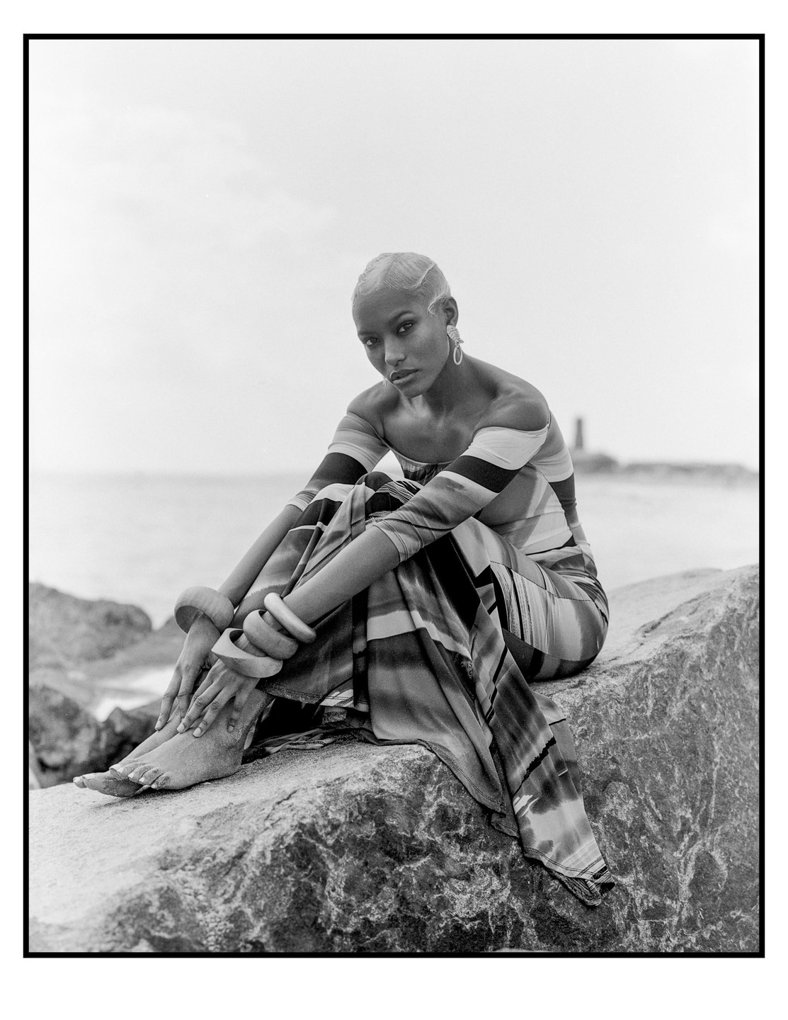 A woman in a striped dress sits on a rock by the ocean.