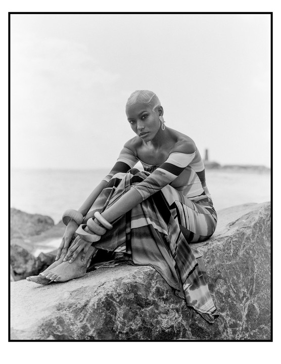 A woman in a striped dress sits on a rock by the ocean.