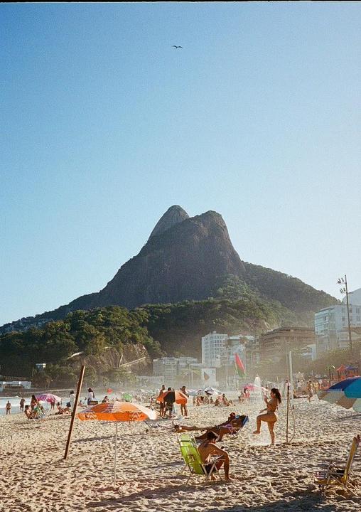 A beach with many people and umbrellas is in the foreground, with a large mountain and buildings in the background.