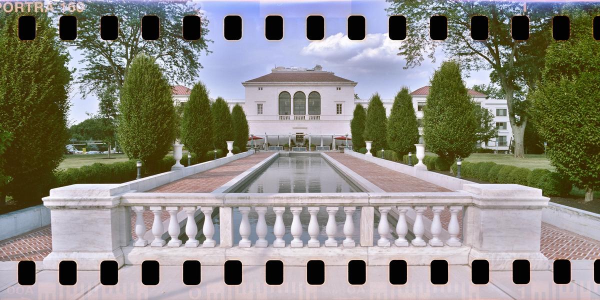 A white mansion with a long reflecting pool and manicured trees is seen from behind a stone balustrade.