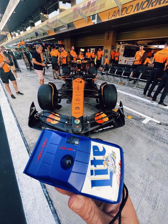 A person holds a blue disposable camera in front of a Formula 1 race car and pit crew.