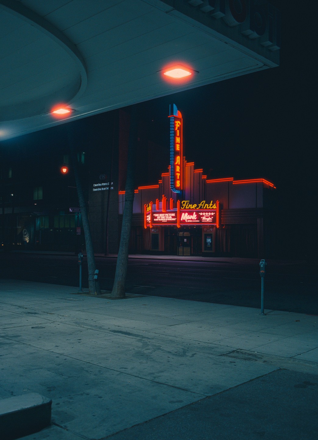 A movie theater with neon signs is visible from a sidewalk at night.