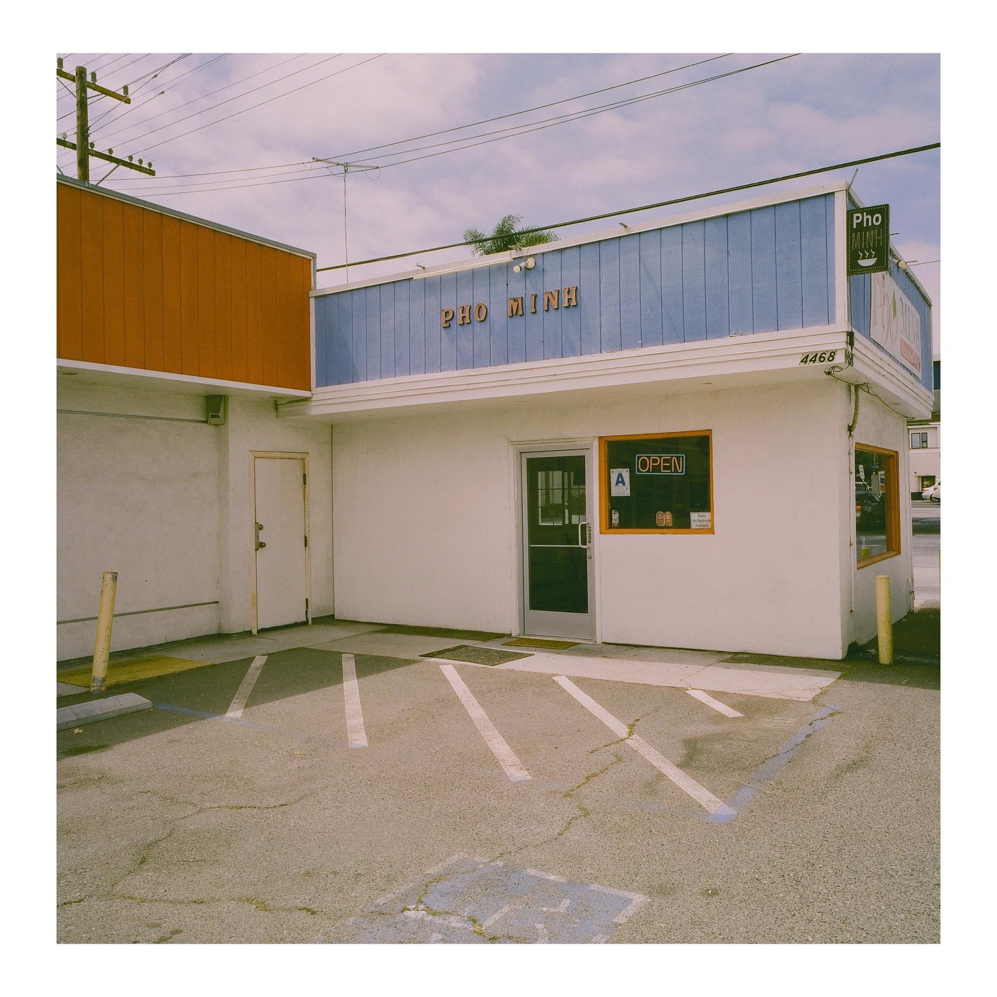 A white building with a blue facade and an orange sign that says "Pho Minh" has a parking lot in front of it.