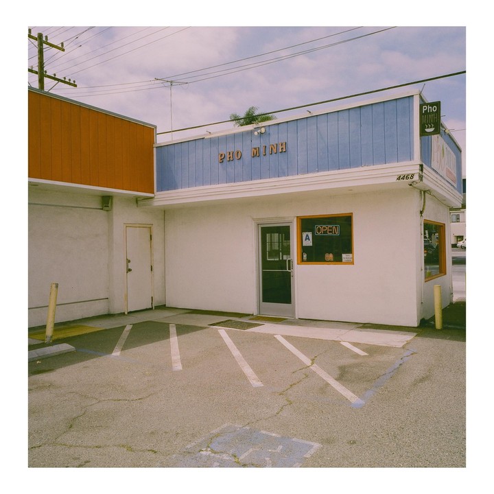 A white building with a blue facade and an orange sign that says "Pho Minh" has a parking lot in front of it.