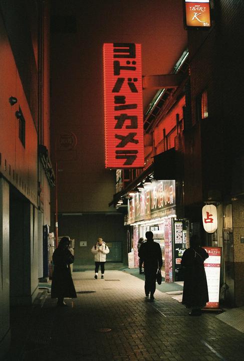 People walk along a narrow street at night, illuminated by a large red neon sign and storefront lights.