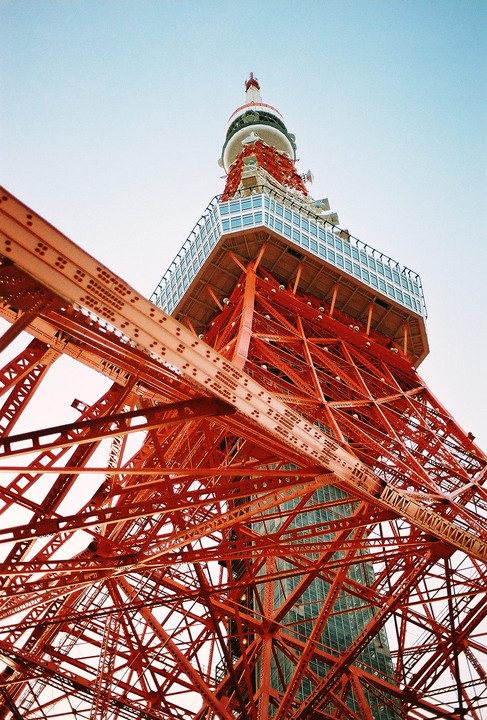 The Tokyo Tower is viewed from below, showing its red metal structure against a light sky.