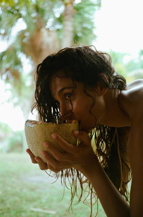 A woman eats a melon outdoors with palm trees in the background.