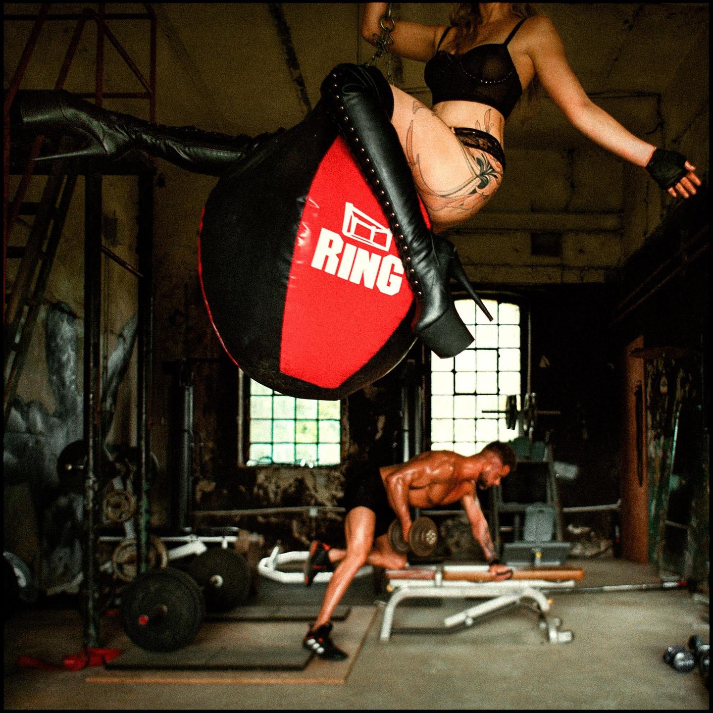 A woman in lingerie and high-heeled boots sits on a punching bag above a man lifting weights in a gym.