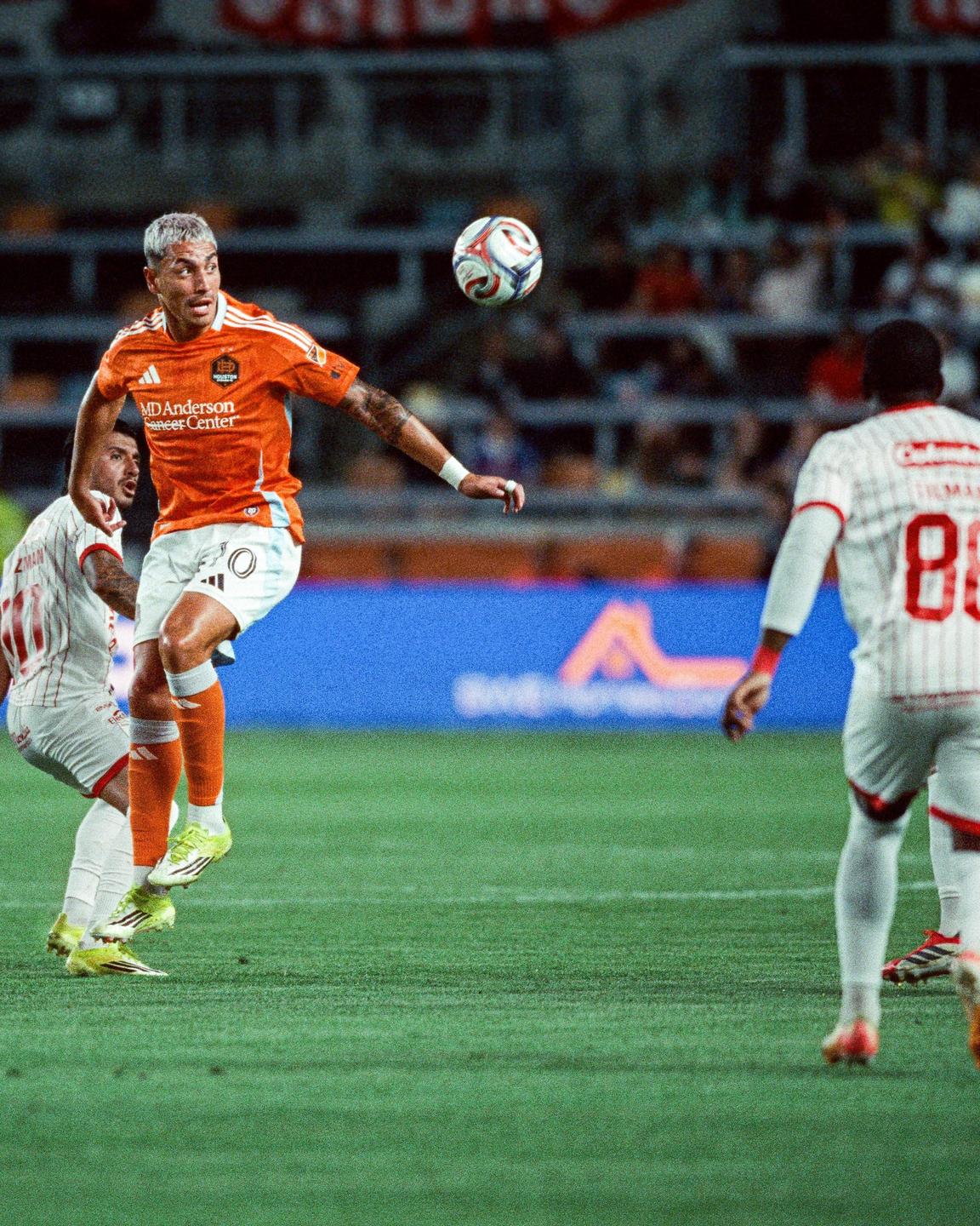 A soccer player in an orange jersey jumps to head a soccer ball during a game.