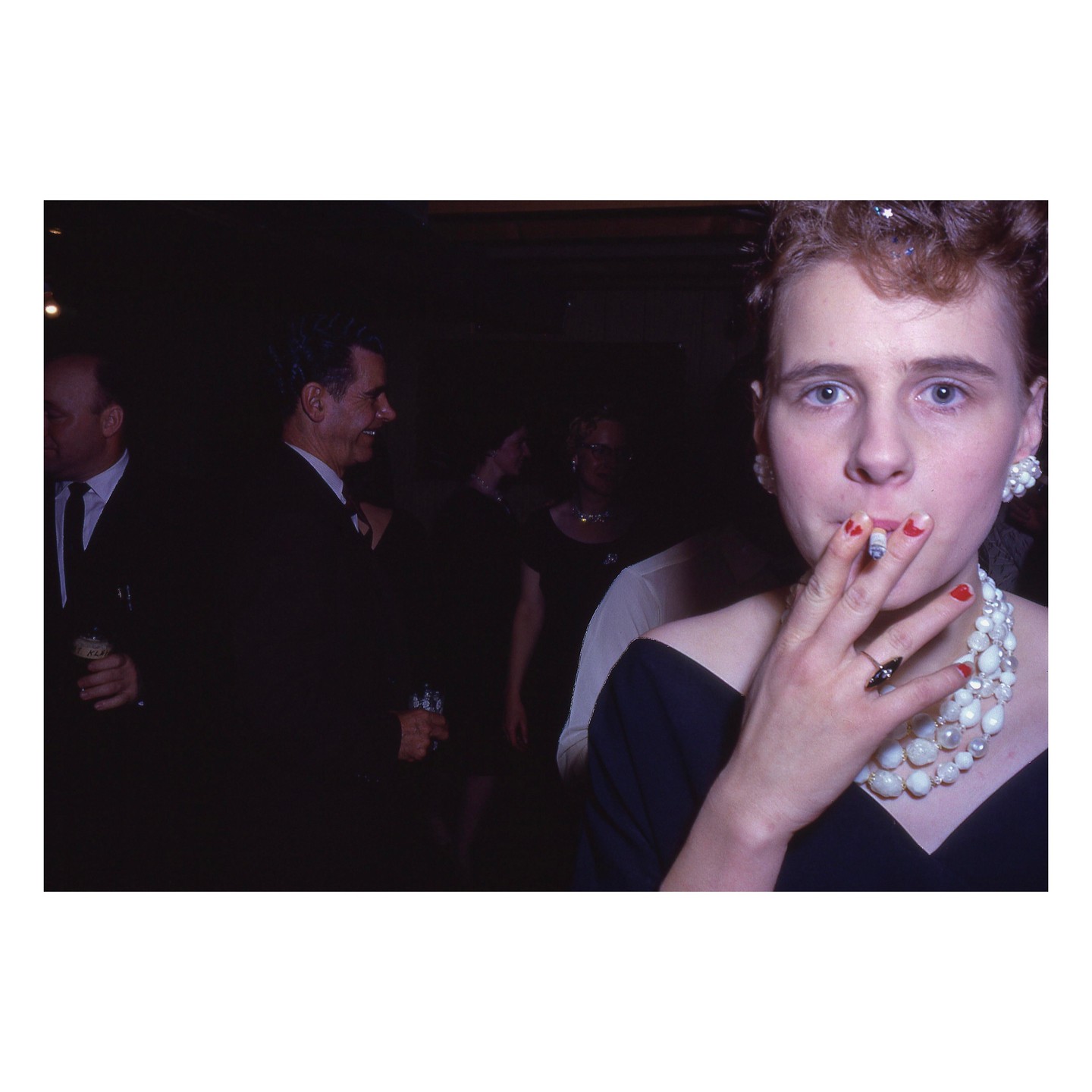 A woman with red fingernails and a pearl necklace smokes a cigarette while looking at the camera, with other people in the background.