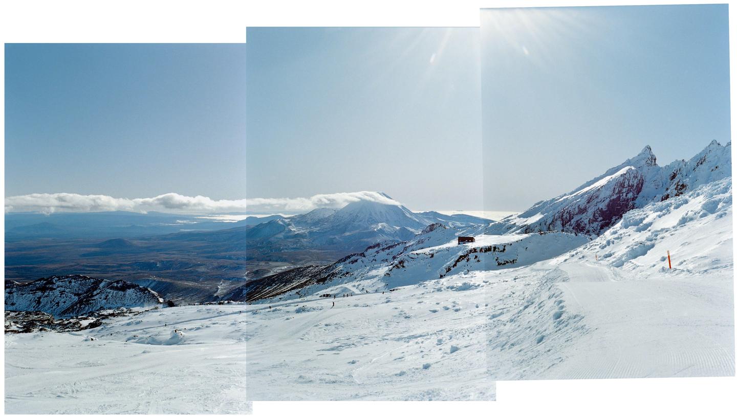 A snow-covered mountain landscape with a small building on a ridge and a distant mountain range under a clear sky.