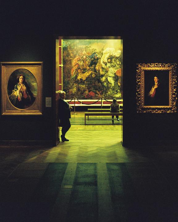 A person stands in a dark museum gallery looking at a large painting, while another person sits on a bench in the background.