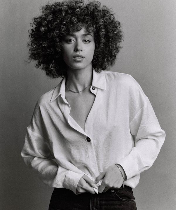 A woman with curly hair wears a white shirt and dark pants while looking at the viewer.