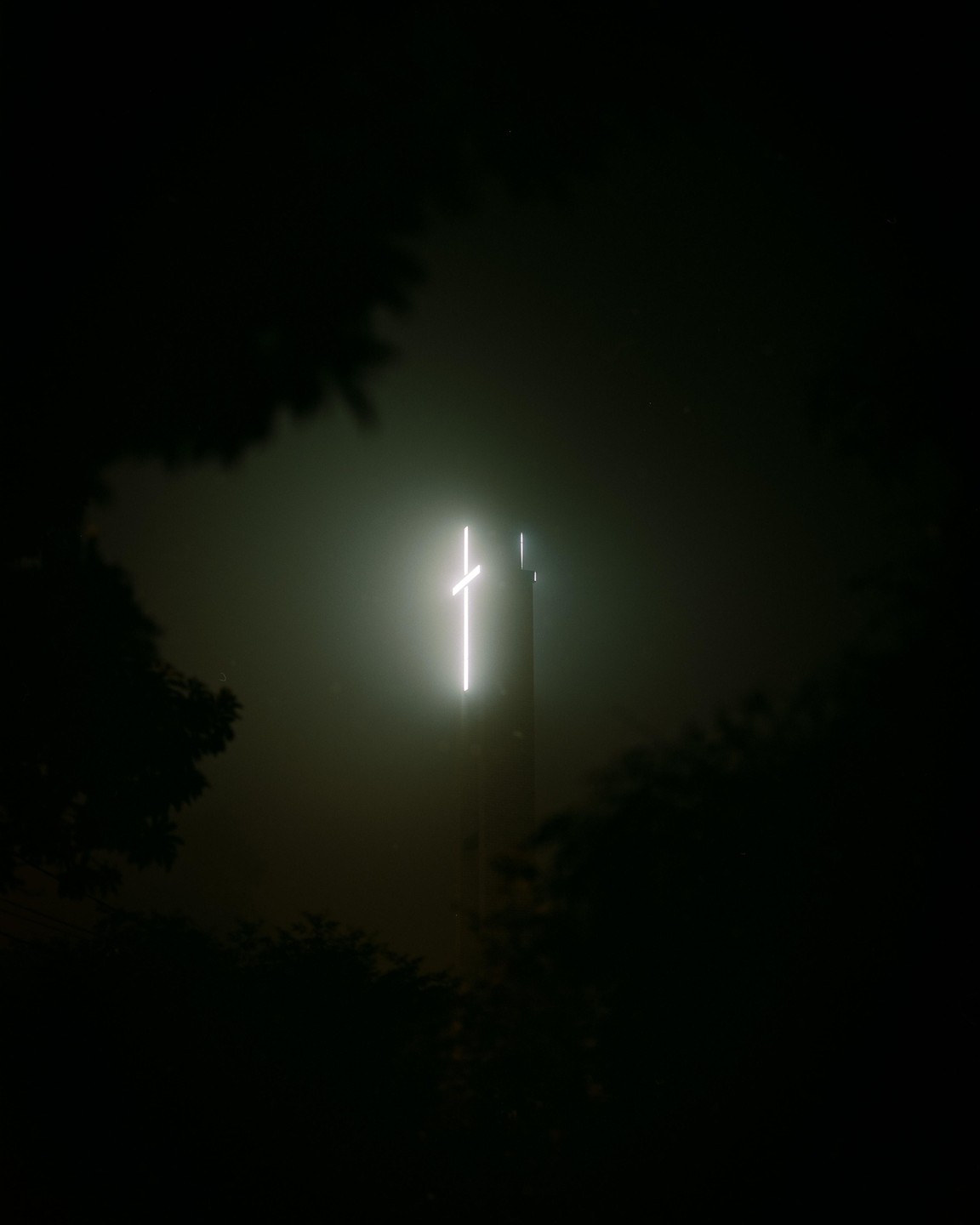A glowing cross is visible on top of a building at night, surrounded by fog and trees.