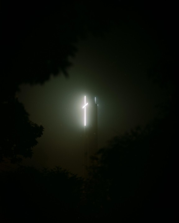 A glowing cross is visible on top of a building at night, surrounded by fog and trees.