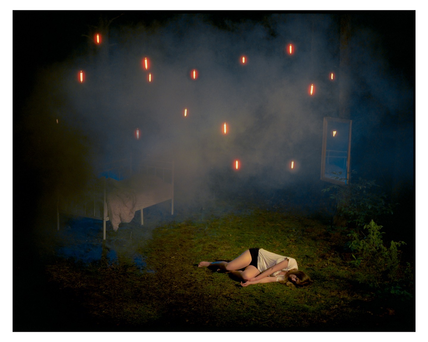 A woman lies on the grass in a hazy forest at night, with a bed and a window frame nearby and glowing orange lights floating above.