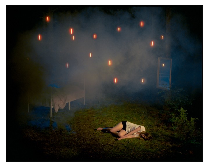 A woman lies on the grass in a hazy forest at night, with a bed and a window frame nearby and glowing orange lights floating above.