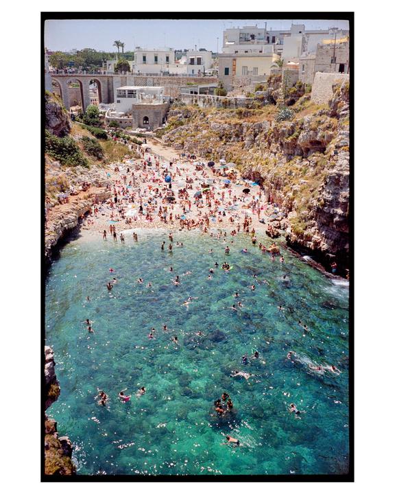 A crowded beach and clear blue water are nestled between cliffs with buildings on top.