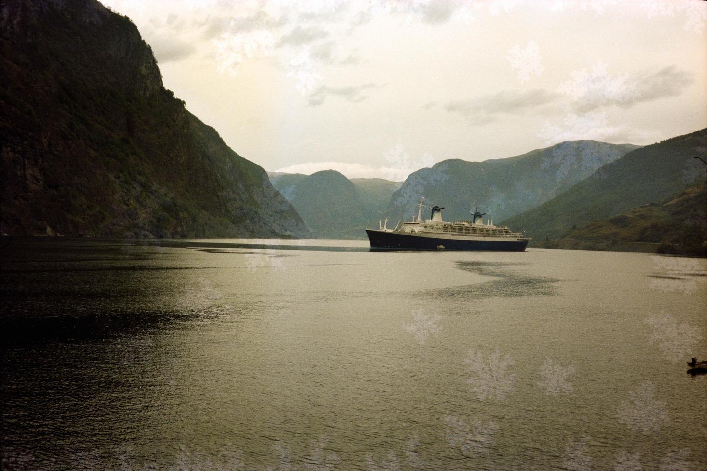 A large ship sails on a body of water surrounded by mountains under a cloudy sky.
