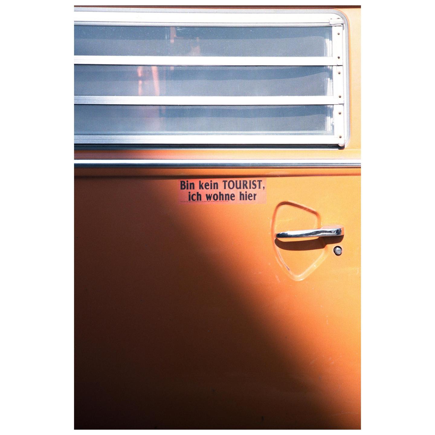 A close-up shows the side of an orange camper van with a window, a door handle, and a sticker that reads "Bin kein TOURIST, ich wohne hier."