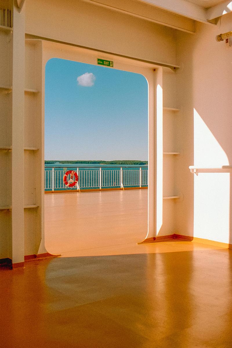 A view from inside a ferry shows the deck, a life preserver, and the sea with a distant shoreline under a blue sky with a single cloud.