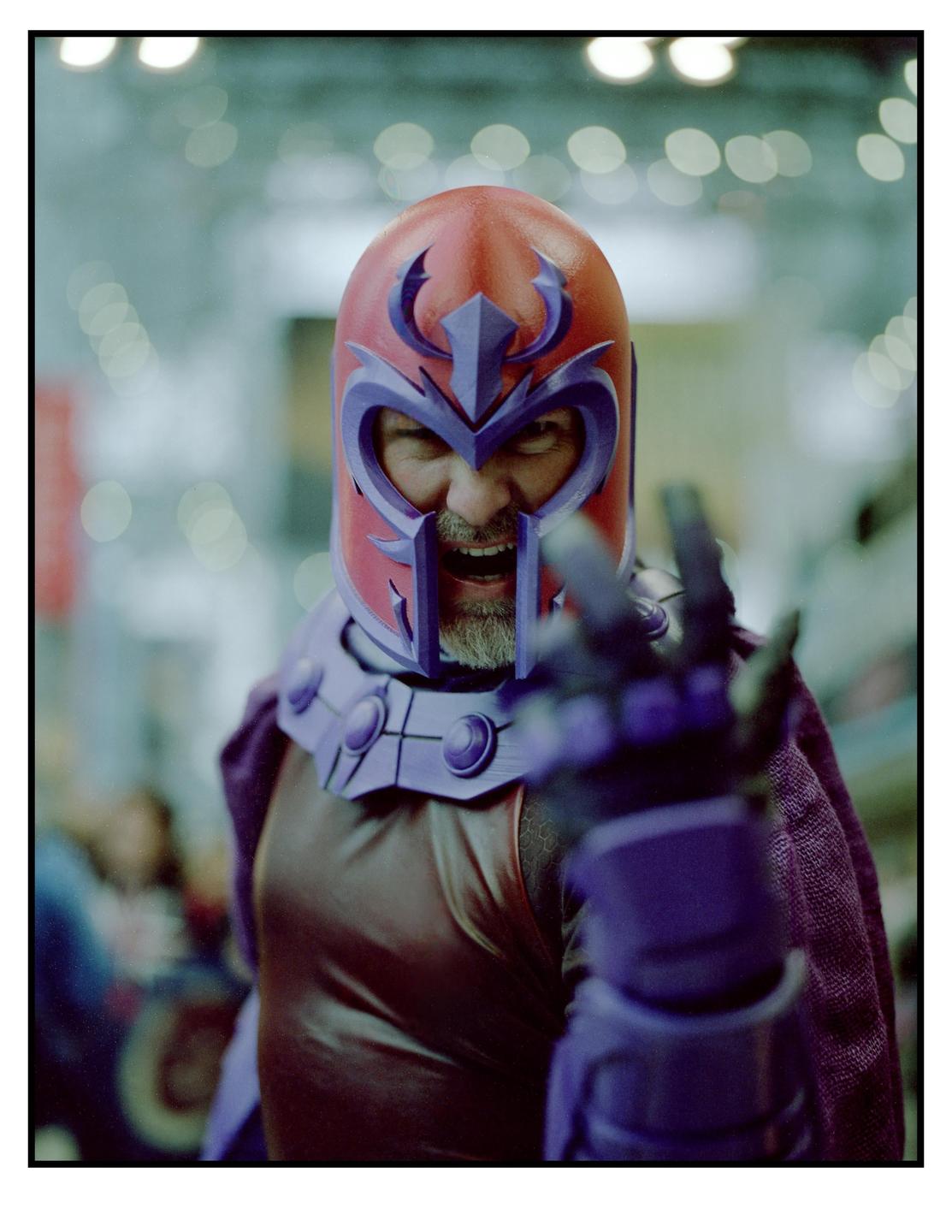 A man in a Magneto costume with a red helmet and purple cape extends his hand towards the viewer.