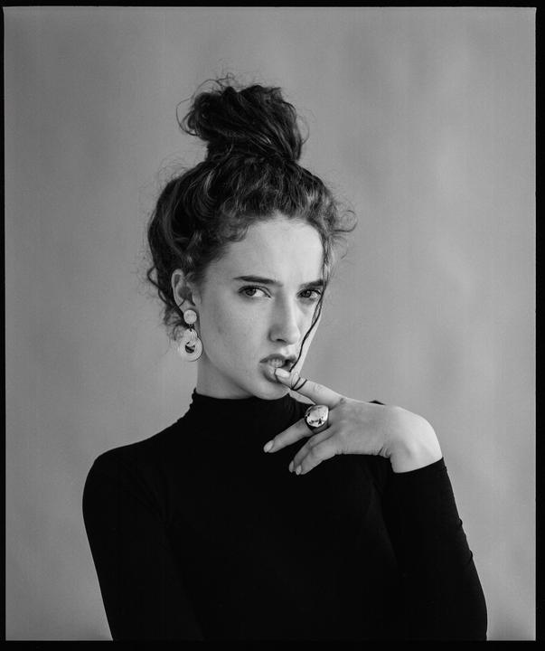 A woman with her hair in a bun and wearing a black turtleneck, earrings, and a ring poses for a portrait.