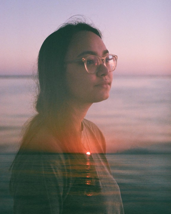 A woman with glasses is shown in a double exposure with a sunset over water.