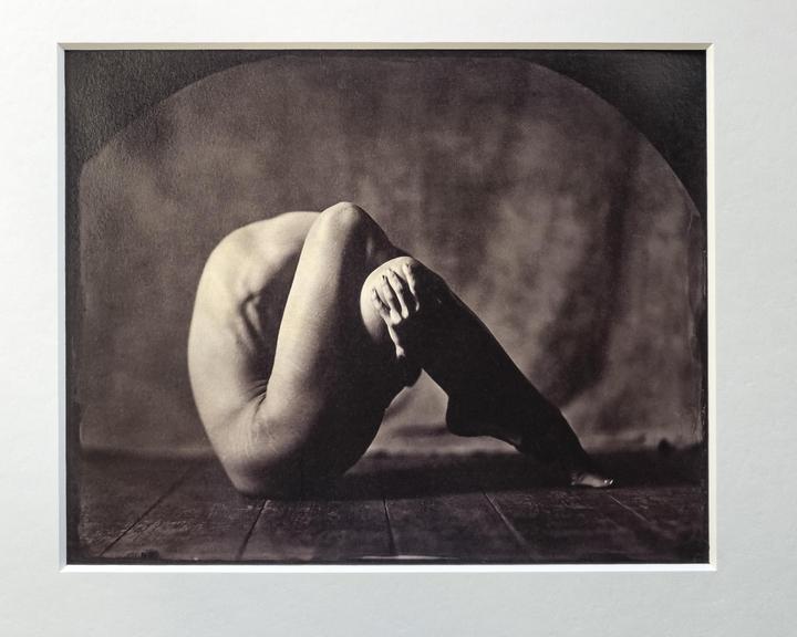 A nude woman is curled up on a wooden floor with her knees bent and her hands clasped around one knee.