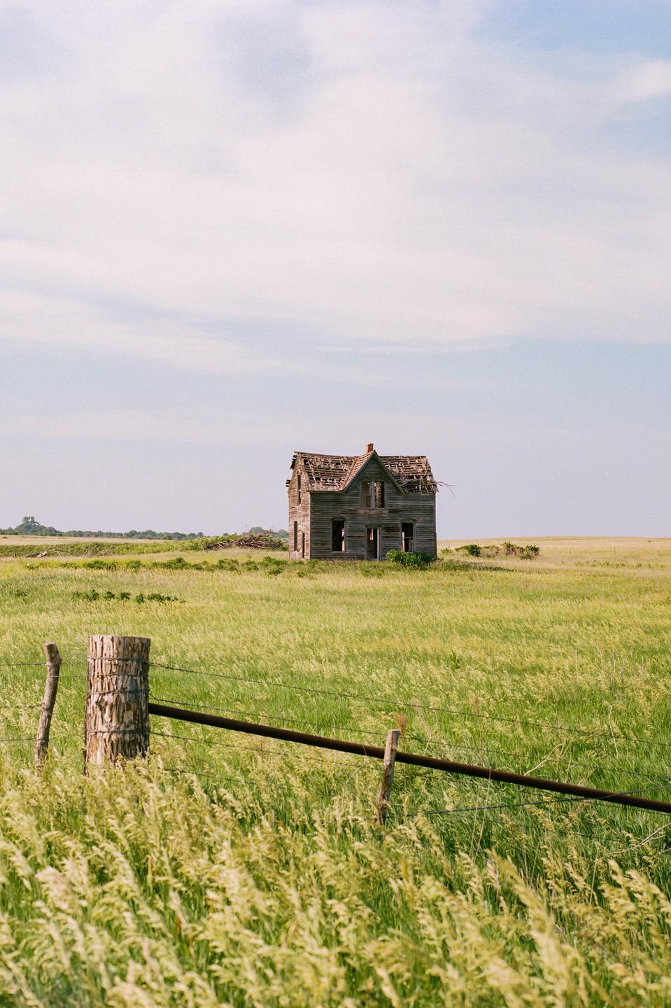An abandoned wooden house with a damaged roof stands in a field of tall grass under a cloudy sky, with a barbed wire fence in the foreground.