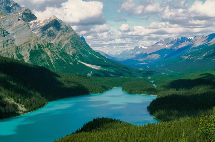 A turquoise lake is surrounded by a dense forest and mountains under a cloudy sky.