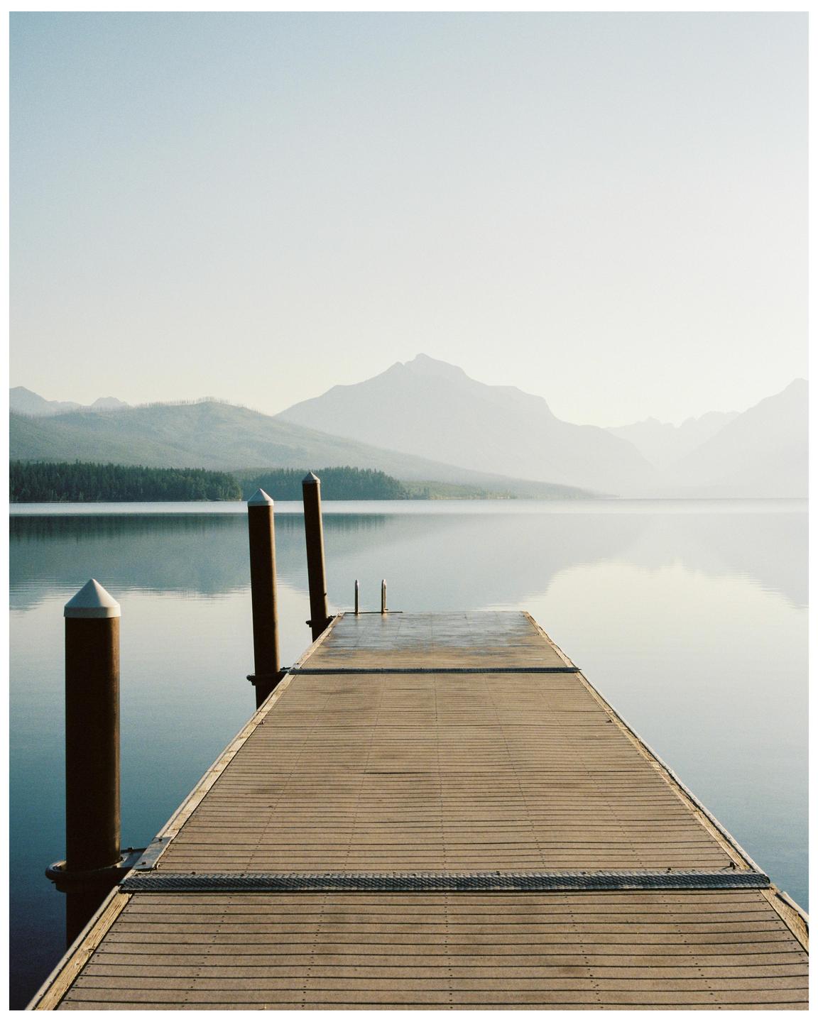 A wooden dock extends into a calm lake with mountains and a hazy sky in the background.