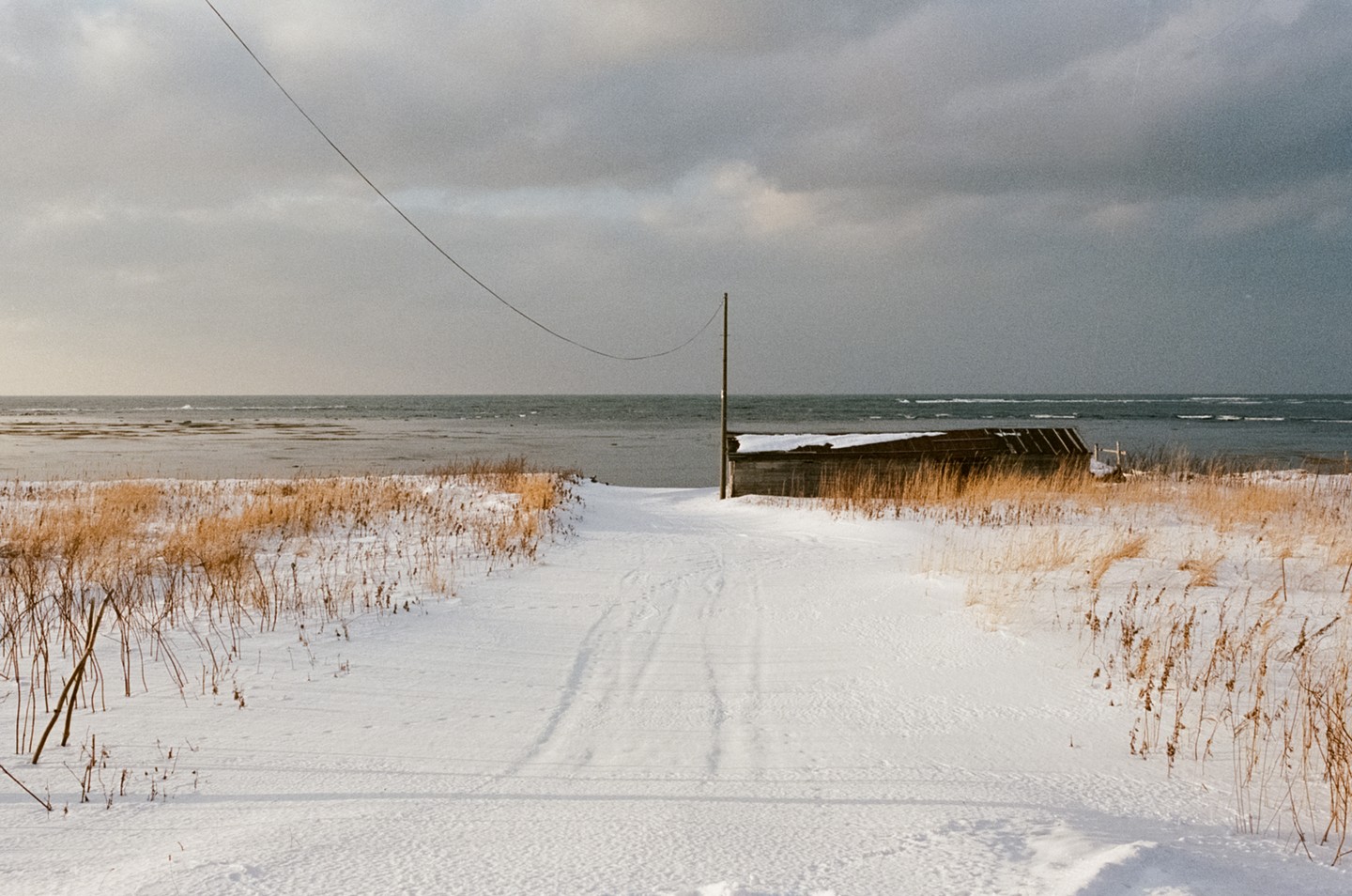 A snow-covered path leads to a body of water with a small structure on the right and a power line stretching across the sky.